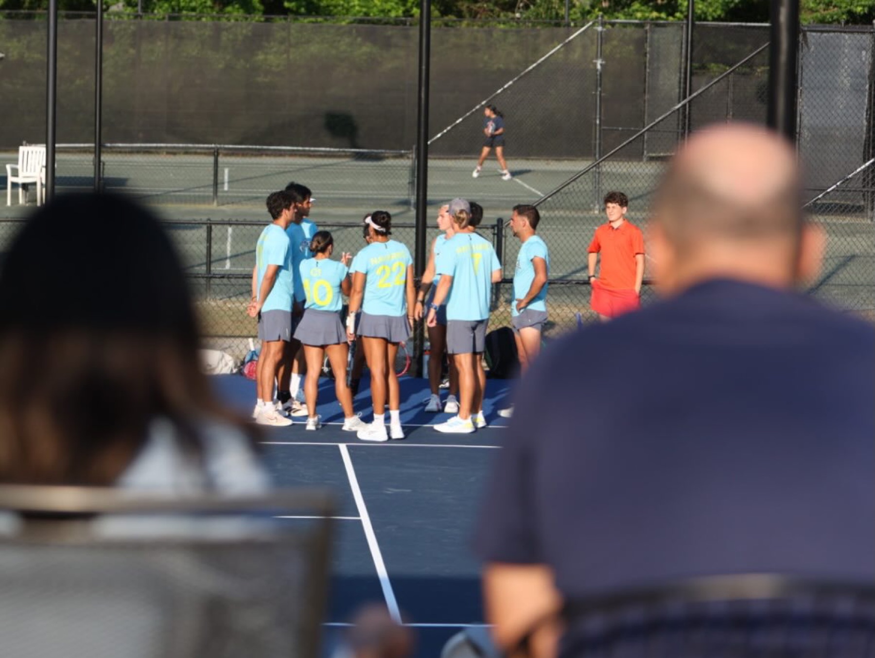 A team huddle on an outdoor court seen over the shoulders of club members watching courtside.