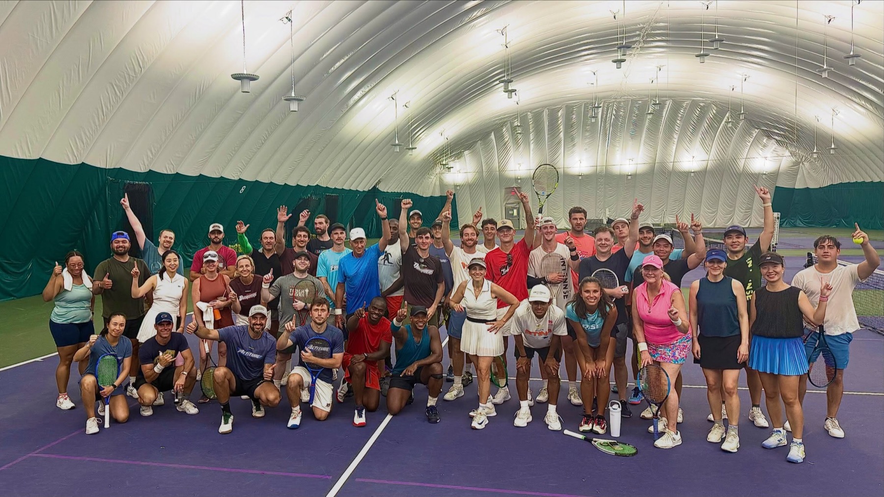 About fifty players of mixed ages gathered on club hard courts inside an inflatable dome, racquets and hands raised.